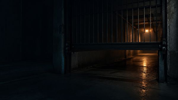 Raskas metalliportti laskettuna puoliväliin pimeän käytävän poikki, pieni lämmin valo takana, rajoituksen tunnelma, limitit