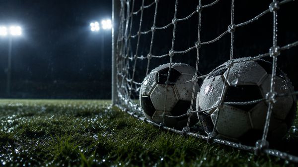 Kaksi jalkapalloa vierekkäin maaliverkossa yöllä, valonheittimet takana, märkä nurmi, lähikuva. BTTS