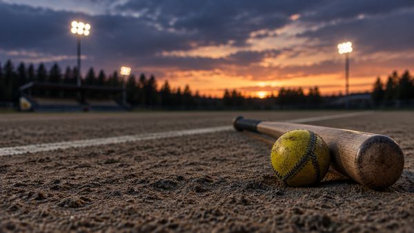 Keltainen pesäpallo ja puinen maila hiekkakentällä iltahämärässä, pienen stadionin valot sumeina, pesäpallo-vedonlyönti