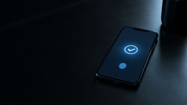 Älypuhelin tummalla pöydällä, näytöllä tyhjä hehkuva maksun hyväksymisnäkymä sormenjälkikuvakkeella, minimalistinen, viileä valo. Apple Pay