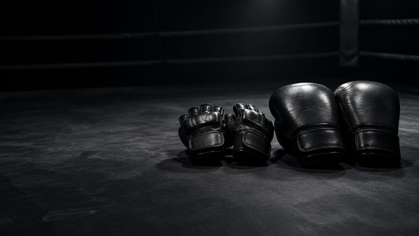 Kaksi MMA-hanskaa ja pari nyrkkeilyhanskoja vierekkäin tummalla kehän kanvaasilattialla, yksi kohdevalo. MMA vs nyrkkeily